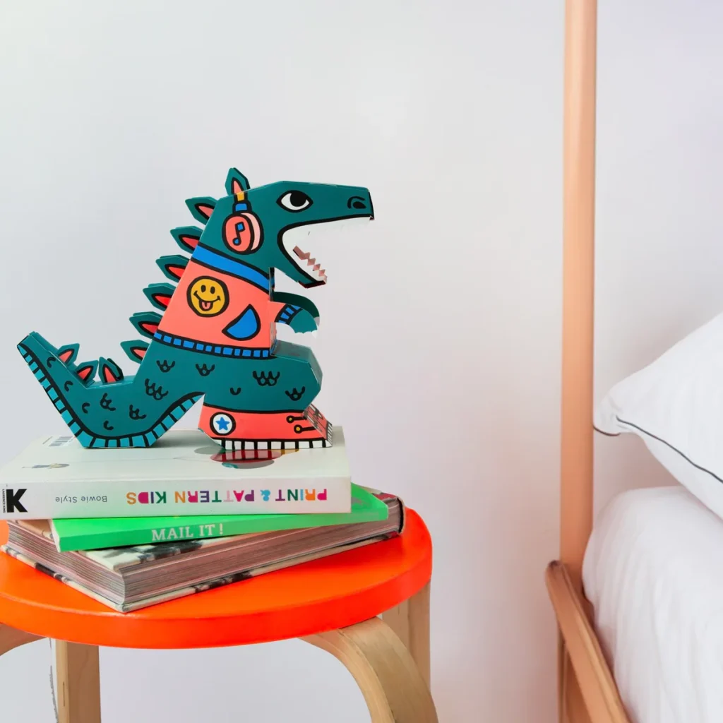 A 3D cardboard dinosaur LED lamp sitting on some books next to a bed