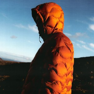 A man is standing in the hills of Scotland wearing a down coat in burnt orange