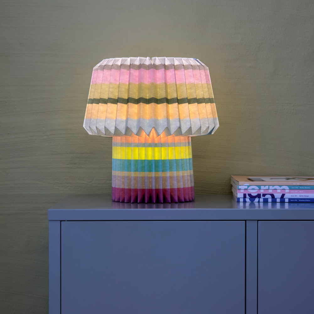 A colorful paper lamp with stripes of bright colors sits on a dresser next to some books