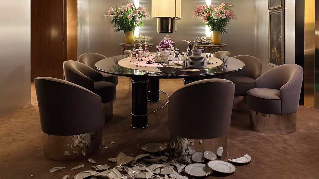 A round glass dining table, 8 dining chairs, broken dishes on the floor, flowers on stands, a chandelier