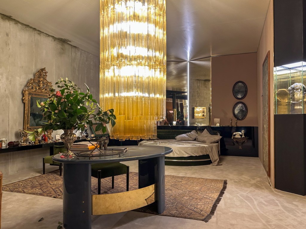 A bedroom set designed by Dimorestudio features a round bed, an oval desk, flowers, and a very large chandelier