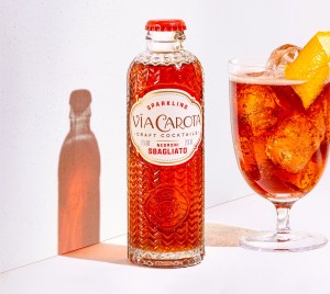 A bottle of Via Carota's Negroni Sbagliato next to a a glass with the cocktail and a lemon twist