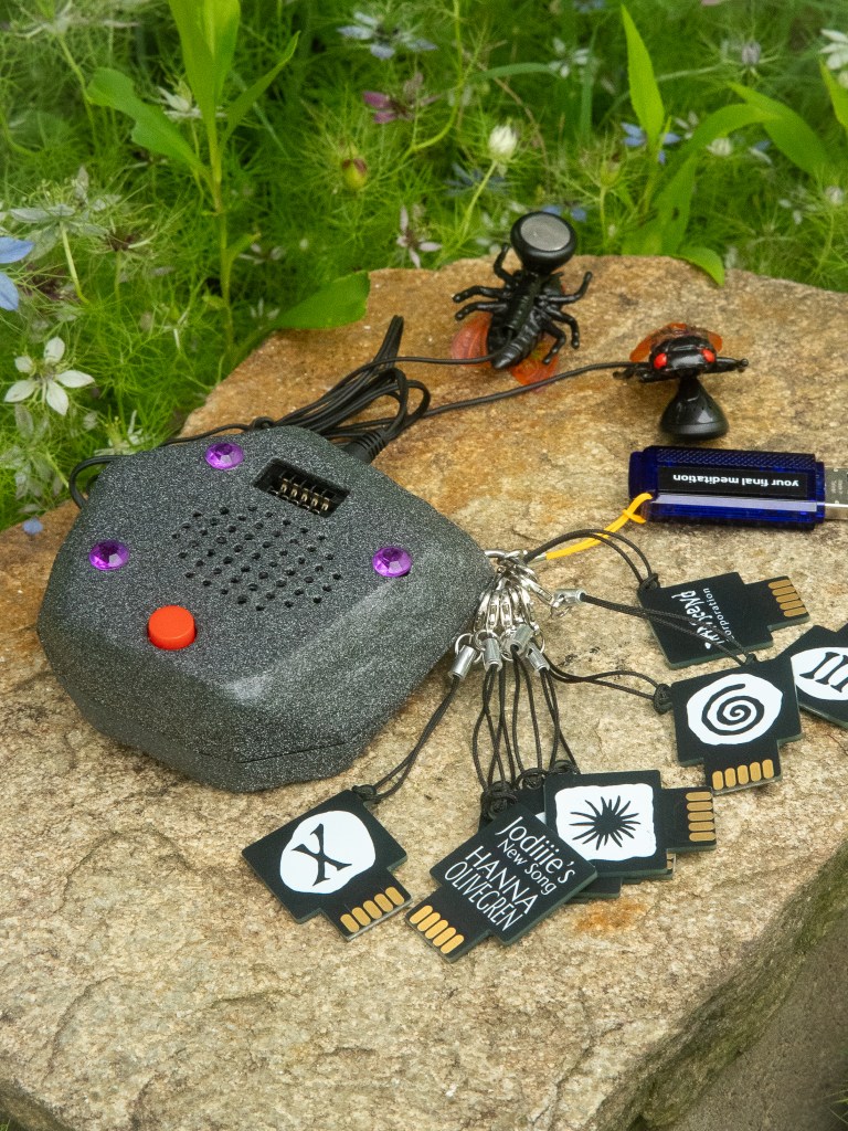 tan rock background with a gray rock-shaped speaker with red and purple buttons with black and white cartridges attached to it