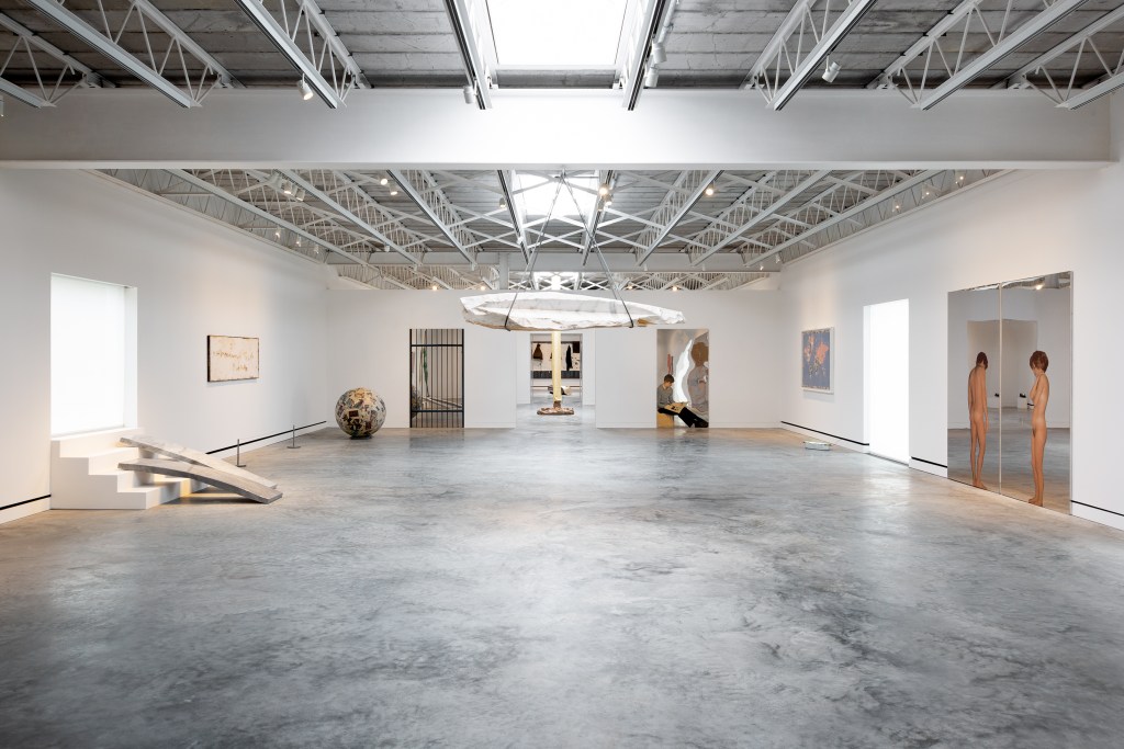 A white walled museum gallery room with concrete floors and several pieces of hanging art and sculptures.