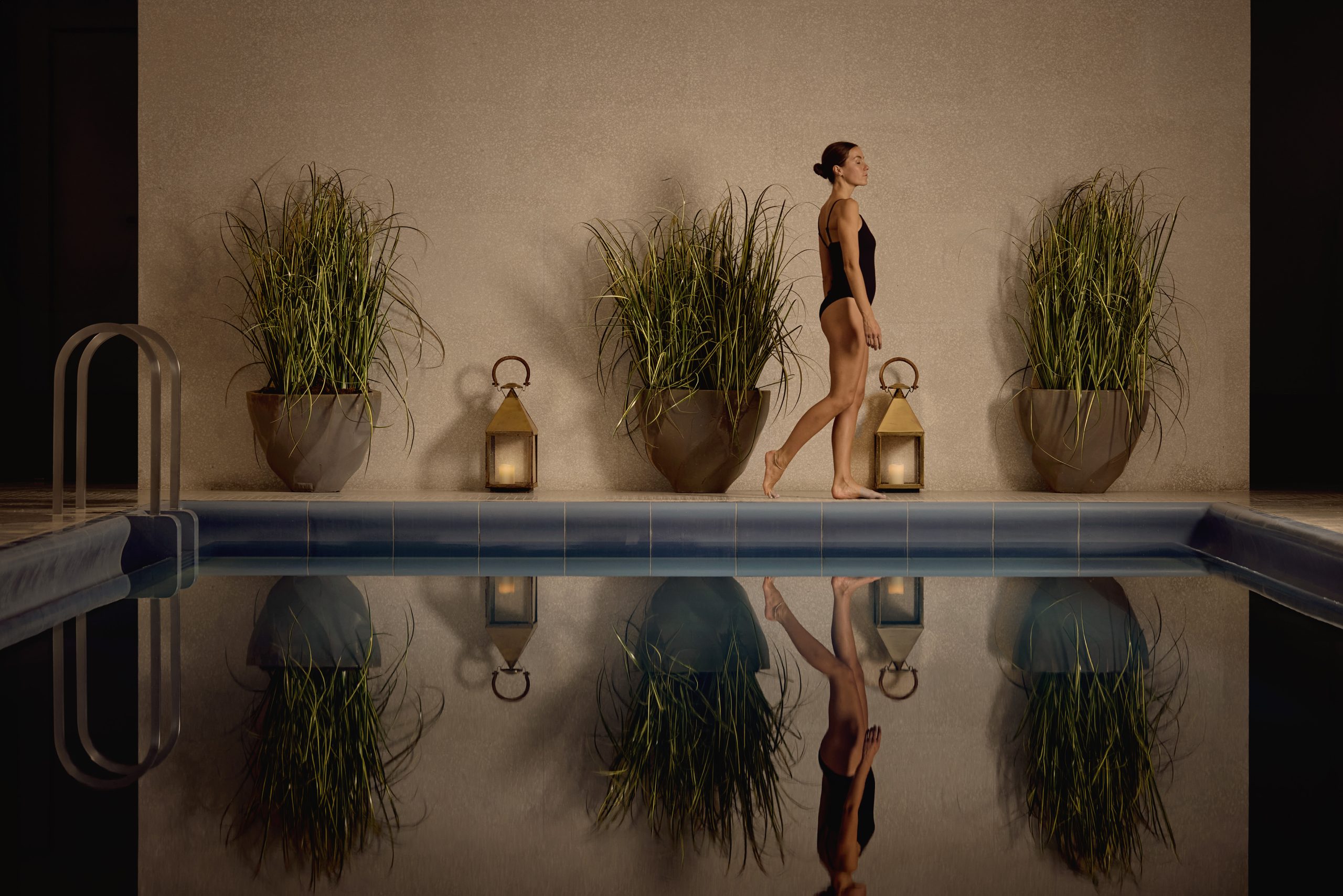 A woman walking by a swimming pool with three plants.