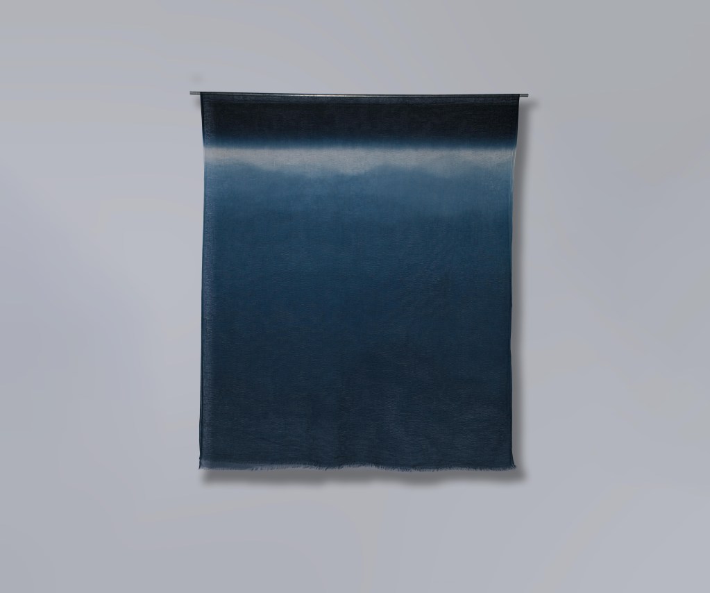 A hand dyed textile hanging on a gray wall.