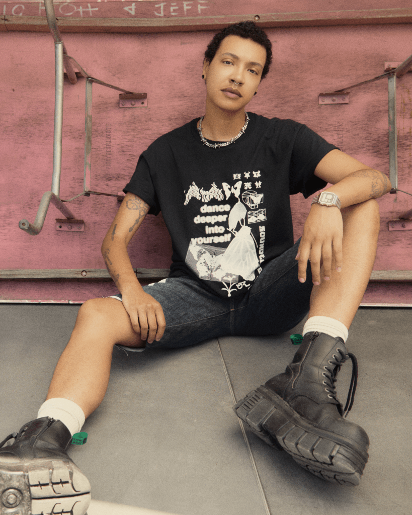 A person wearing an Infinite Transition t-shirt while sitting on the ground and leaning against a pink wall. 