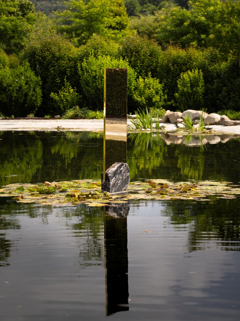 A tall, column-like reflective sculpture in the middle of a pond, surrounded by lily pads.