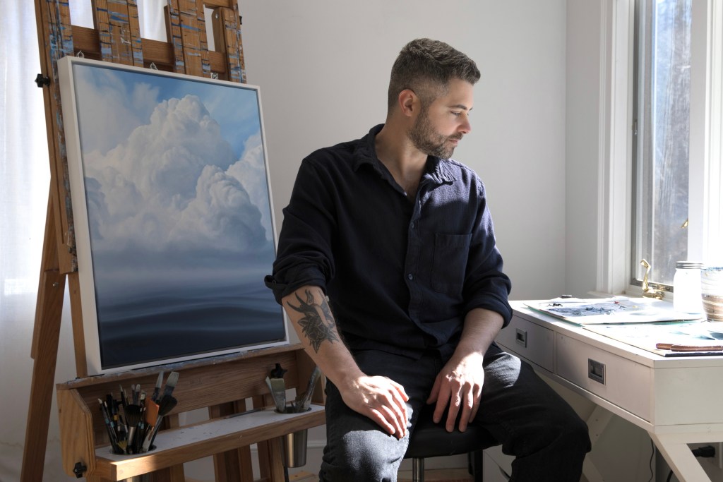 A photo of Derek Macara sitting at his easel, looking down at a desk.