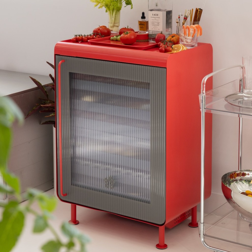 The new limited edition tomato red Rocco smart fridge, shown against a wall, next to a cart, with drinks and accessories on top of it
