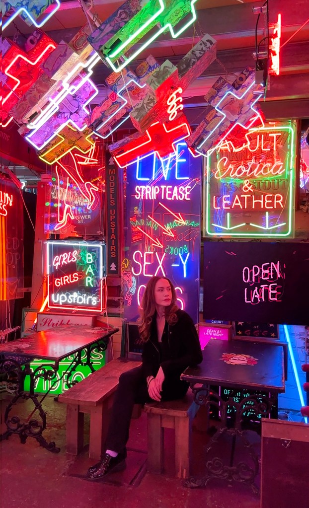 Katherine McMahon seated in a room full of neon signs, next to her neon sign paining that says Girls, Girls, Girls.