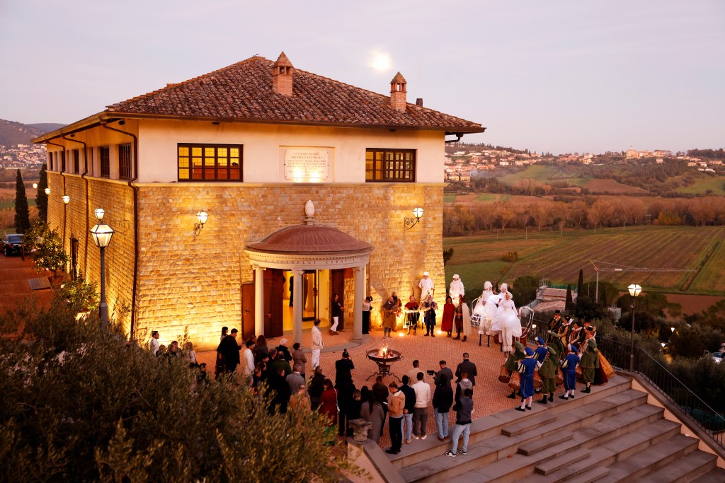 A group of people, including people wearing historic costumes, gather outside Brunello Cucinelli's winery at dusk.