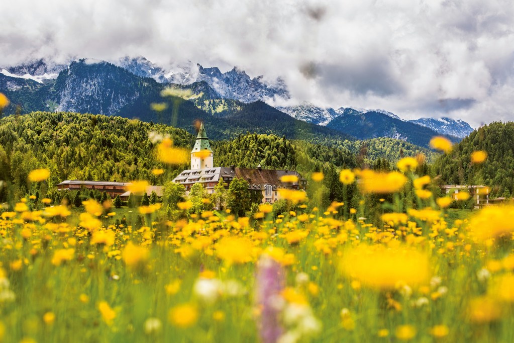 An exterior shot of the Hideaway at Schloss Elmau with yellow wildflowers in the foreground and the Alps in the background.