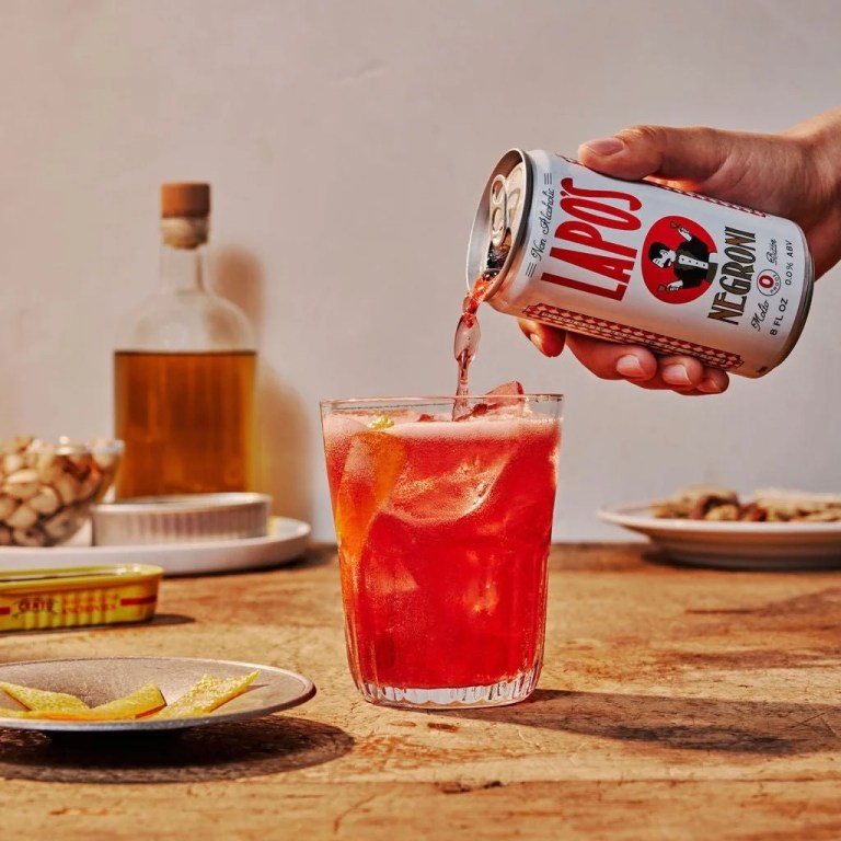 A hand holds a can of Lapo's non-alcoholic Negroni beverage and it is being poured into a clear glass with ice and an orange zest, with other items on the table in the background