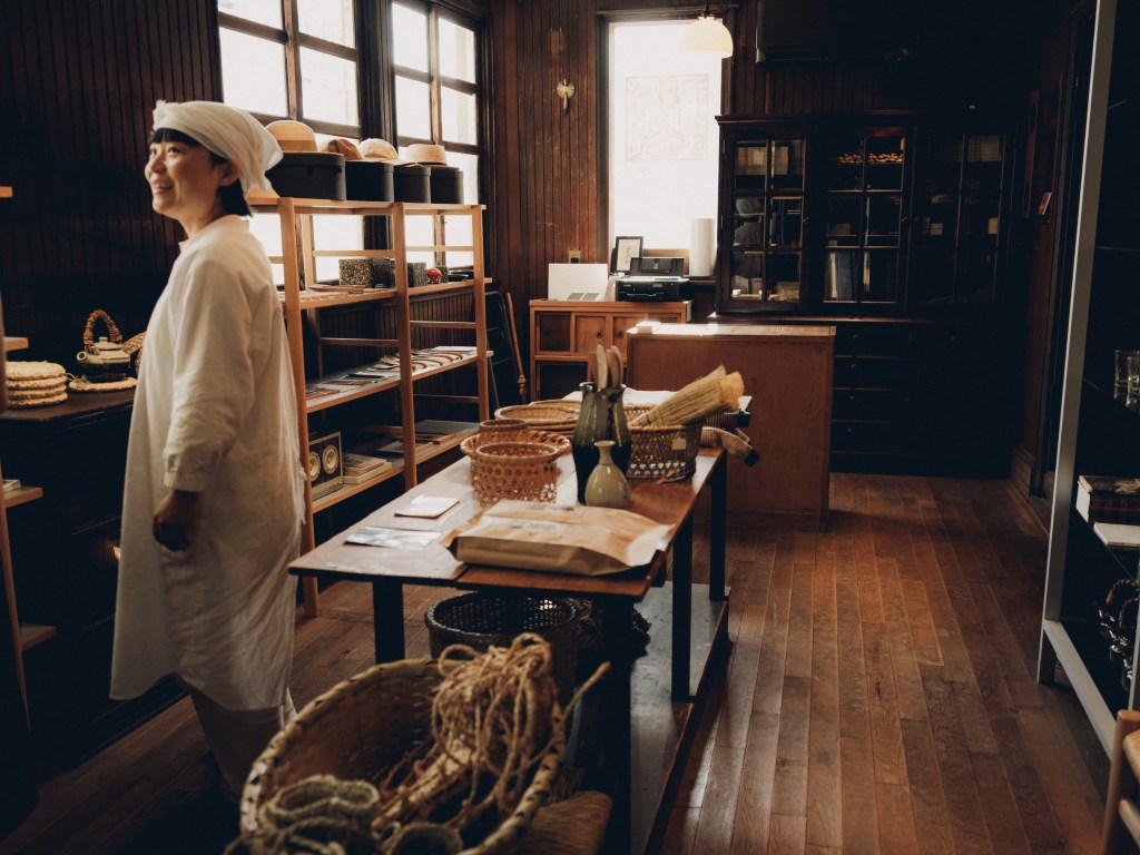 A woman standing in her atelier, surrounded by woven objects for sale.