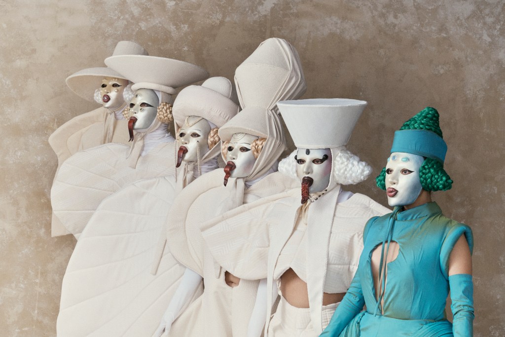 Six performers in Orly Anan-designed costumes, standing in a staggered row.