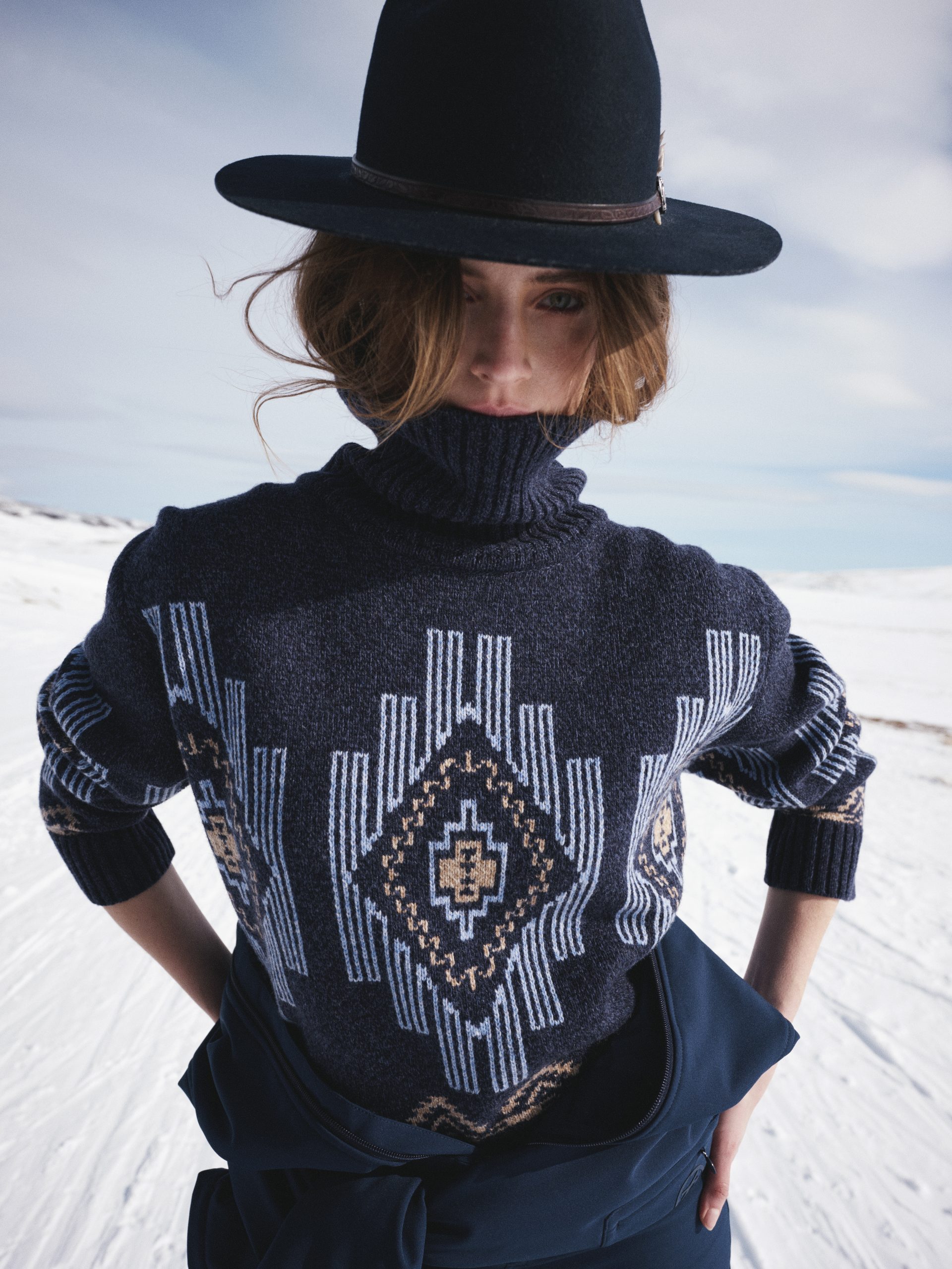 A model with a patterned sweater and a hat with snow and sky in the background.