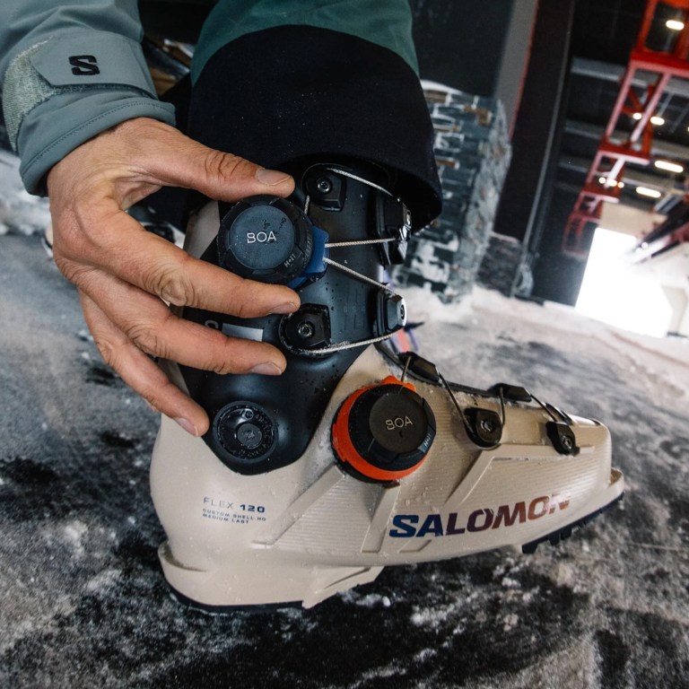 Salomon's S/Pro Supra Ual Boa 120 ski boot shown in a light color with orange and black details, and string-based tensioners shown on the ground outside with a man's hand adjusting a dial
