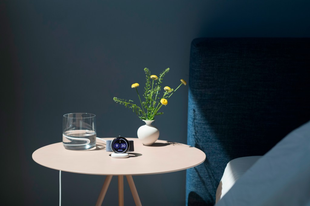 A Google Pixel Watch 4 charging on a bedside table next to a glass of water and a white vase of yellow flowers.