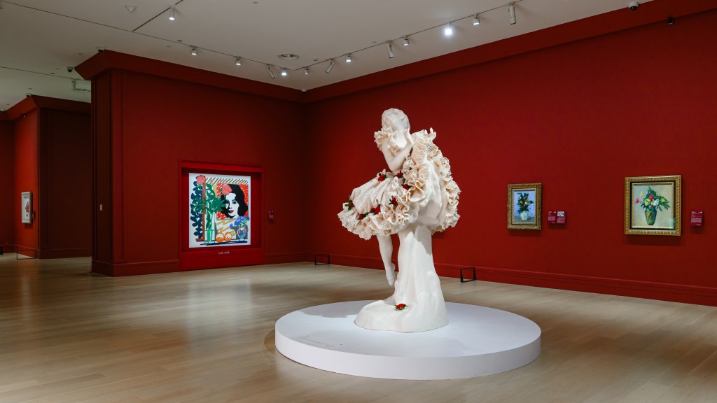 A Jeff Koons ballerina sculpture is displayed in the middle of a red gallery room at Deji Art Museum, surrounded by framed floral art works.