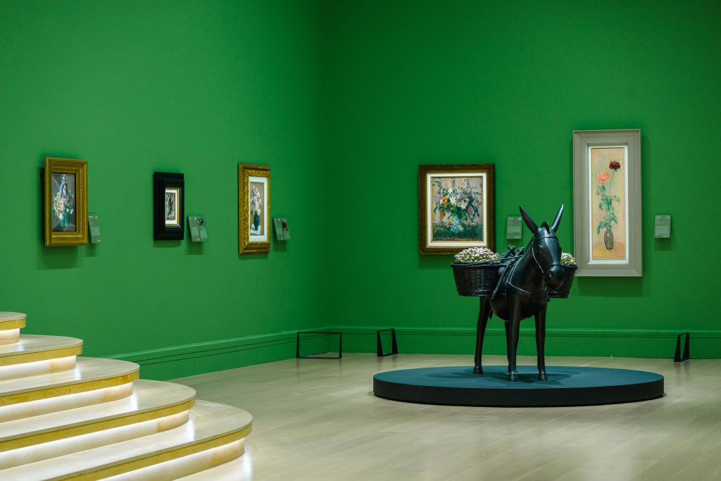 A green room in the Deji Art Museum with stairs on the left and a donkey sculpture in the middle, surrounded by framed floral works of art.