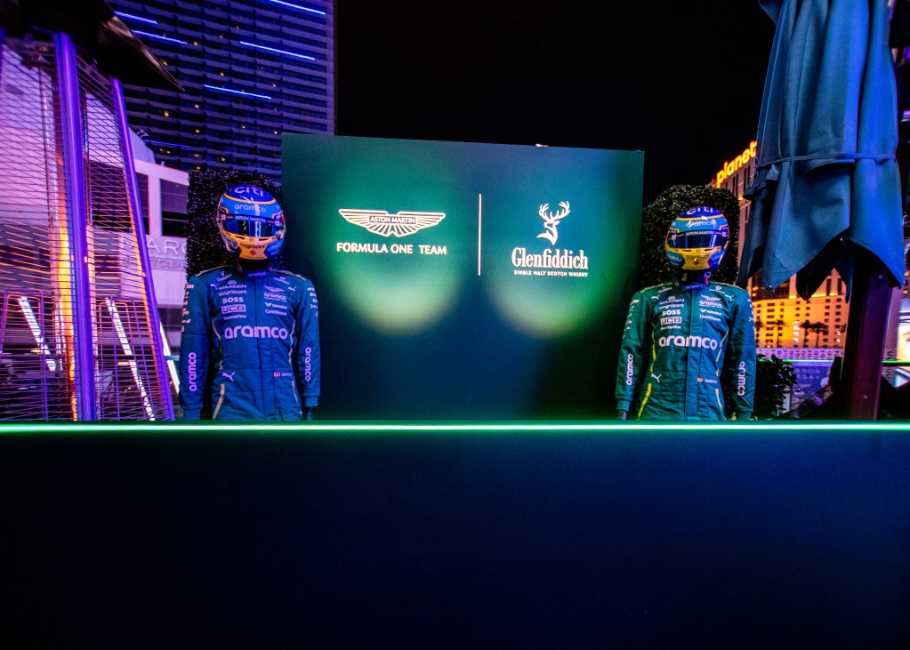 Two people wearing an Aston Martin F1 uniform, flanking an Aston Martin Formula 1 Team and Glenfiddich sign.