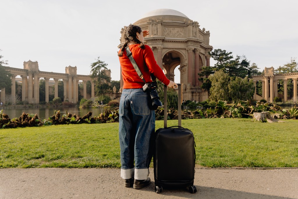 A woman wearing a red sweater looking at a park in San Francisco with a Peak Design roller bag to her right side.