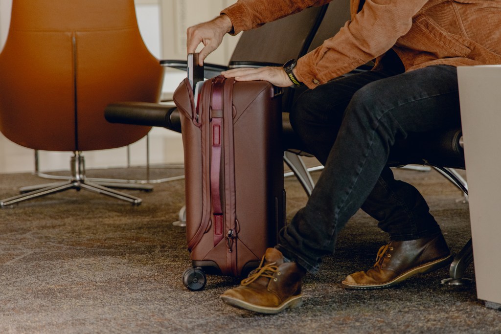 A man sitting in an airport with a Peak Design Roller Pro bag show from the side, showing its zipper and integrated laptop sleeve.