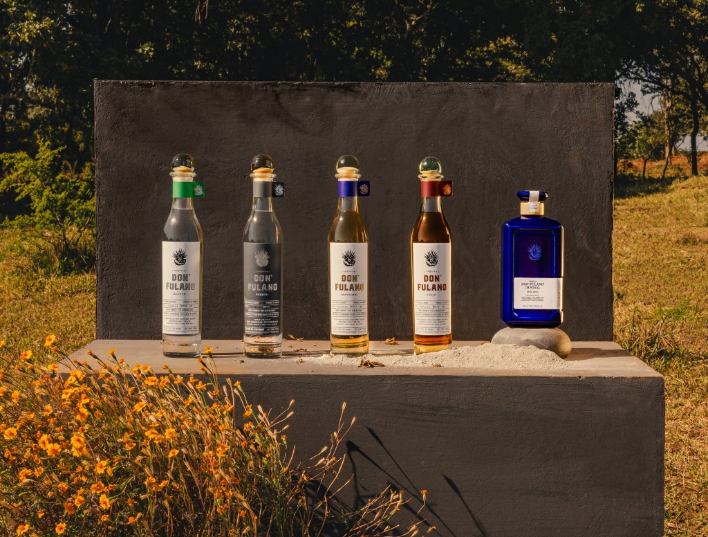 A row of five different varietals of Don Fulano tequila on a table with a gray slate background and yellow flowers in the foreground.