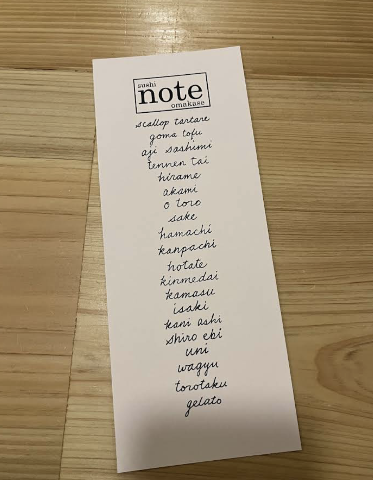 A hand-written vertical paper menu that says "Sushi Note Omakase" at the top of the paper.