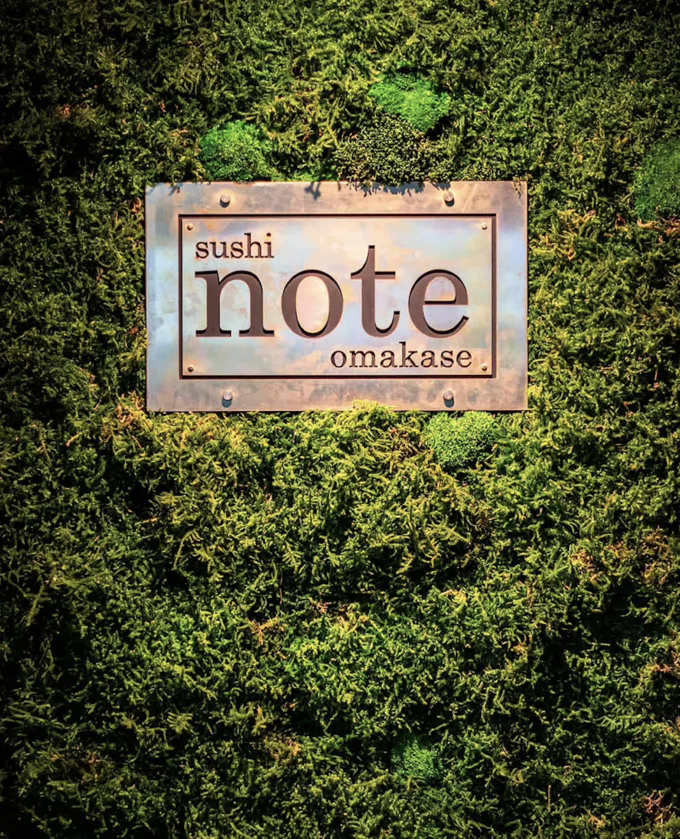 A sign that says "Sushi Note Omakase," hanging on a green plant wall.