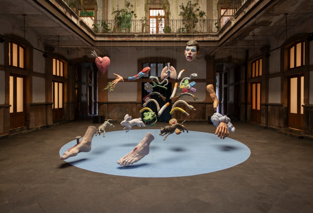 A 3D printed sculpture by Enrique López Llamas, with a clown head with animals and objects that comprise the body glows in the natural light filtering down from above.