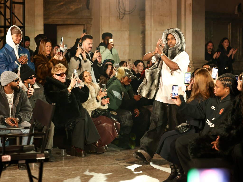 A$AP Rocky wearing a fur hood and raising his hands in a prayer position while people surrounding him take photos with their phones.