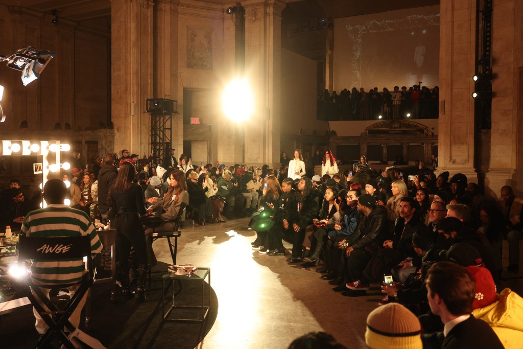 A large, tall room in Manhattan's Financial District with models getting their makeup done in the center and people sitting in rows of chairs watching an AWGE runway show.
