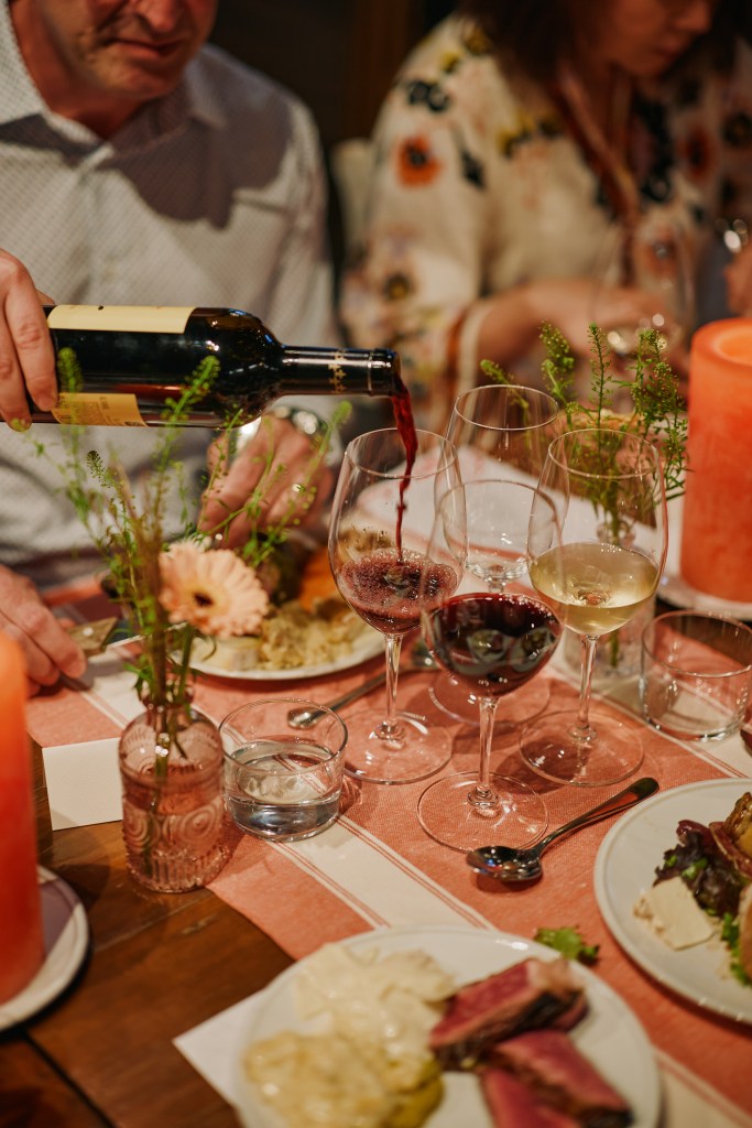 A group in soft focus eating at a table while a server pours red wine into a glass.