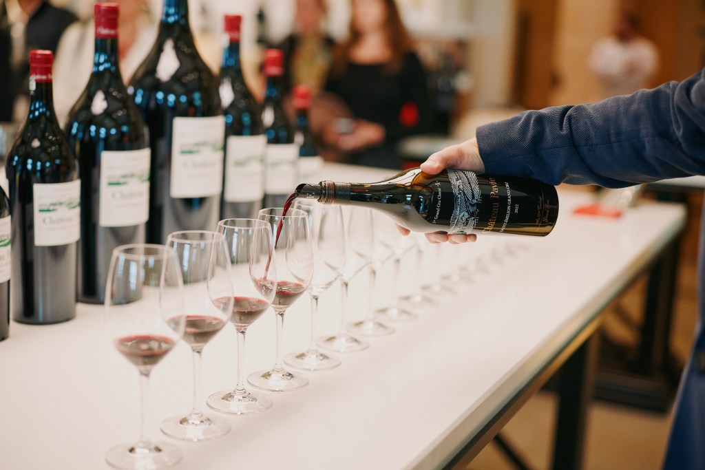 A man's arm pouring tastings of red wine into a row of wineglasses.