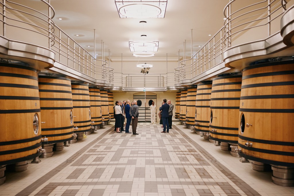 A group of people standing in between rows of large wine barrels in an indoor aging facility.