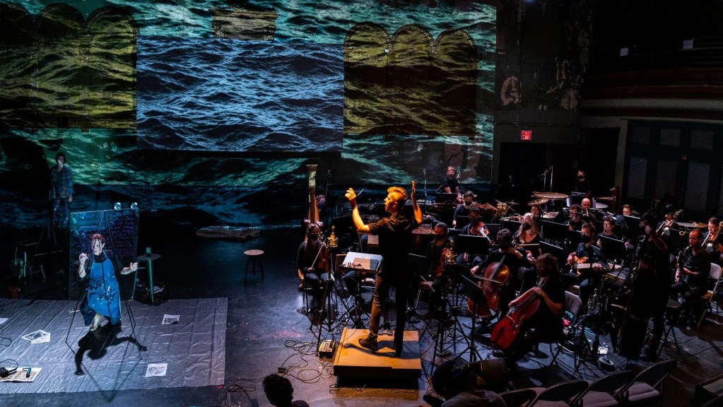A conductor leading an orchestra while a painter paints to the left side of the stage.