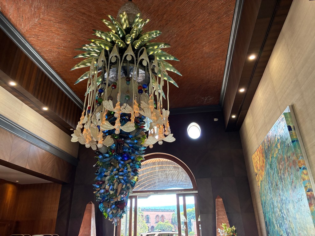 A large glass and wood chandelier depicting bees and agave plants, hanging from a tall, brick-lined ceiling.