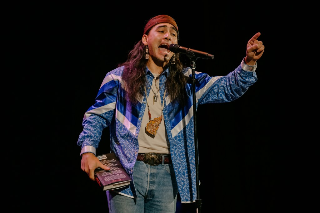 Julian Brave NoiseCat passionately speaking into a microphone while holidng a book in his left hand. 