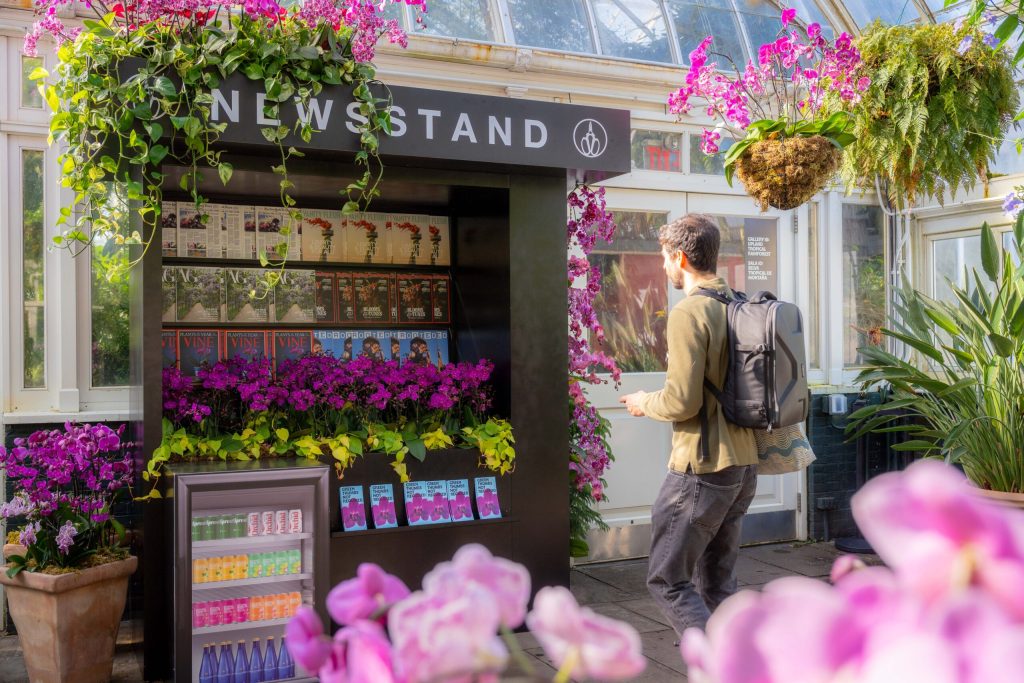 A man wearing a backpack walking towards a newsstand surrounding by colorful flowers with floral-themed magazines on the shelves. 