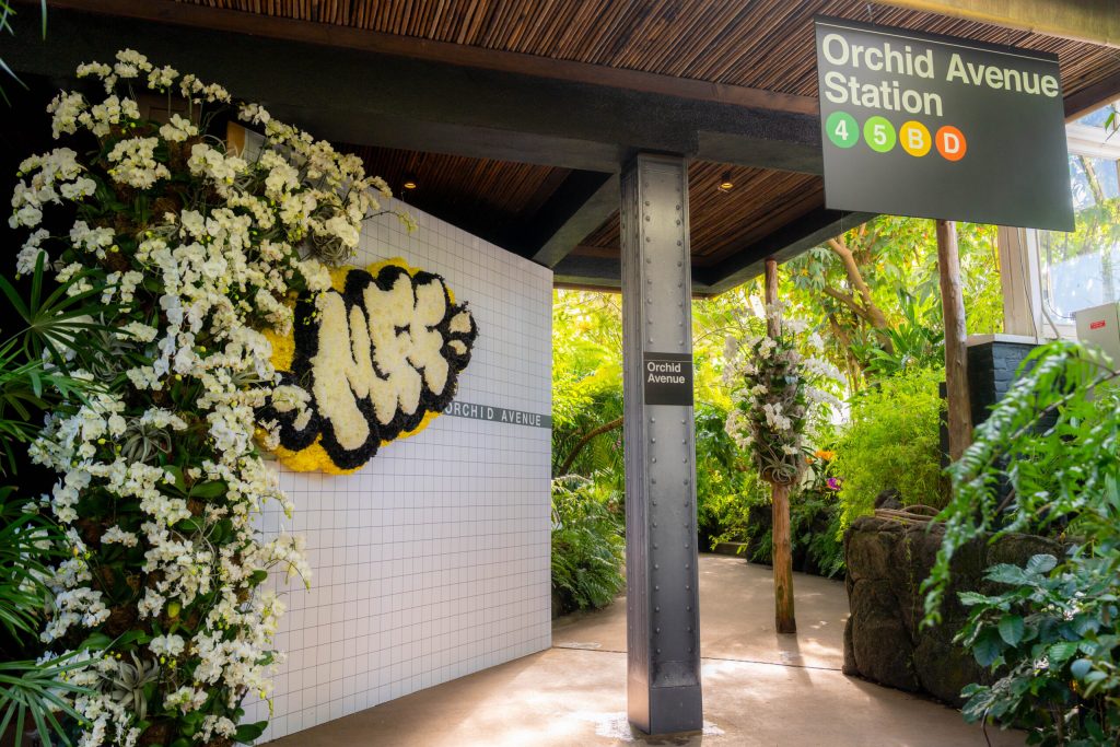 A tiled corner with floral graffiti, a vertical steel beam with an "Orchid Avenue" subway sign and a larger sign hanging from the ceiling that says "Orchid Avenue Station." 
