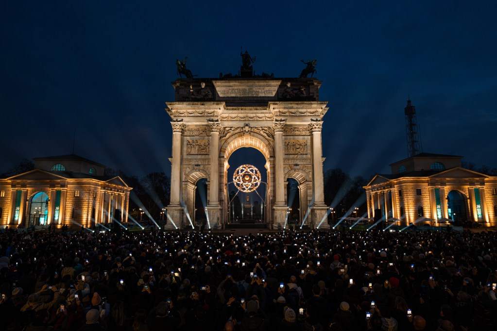 A large crowd of people standing in front of Italy's Arco Della Pace with their phone cameras out, shot at night.