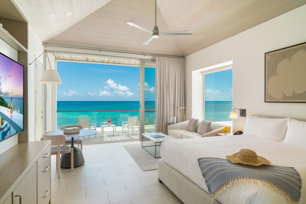 An interior view of an Oceanfront Pool Studio at Rock House with soft white walls and bedding, a loveseat by a small window, and a large sliding door opening to a private patio overlooking the ocean.