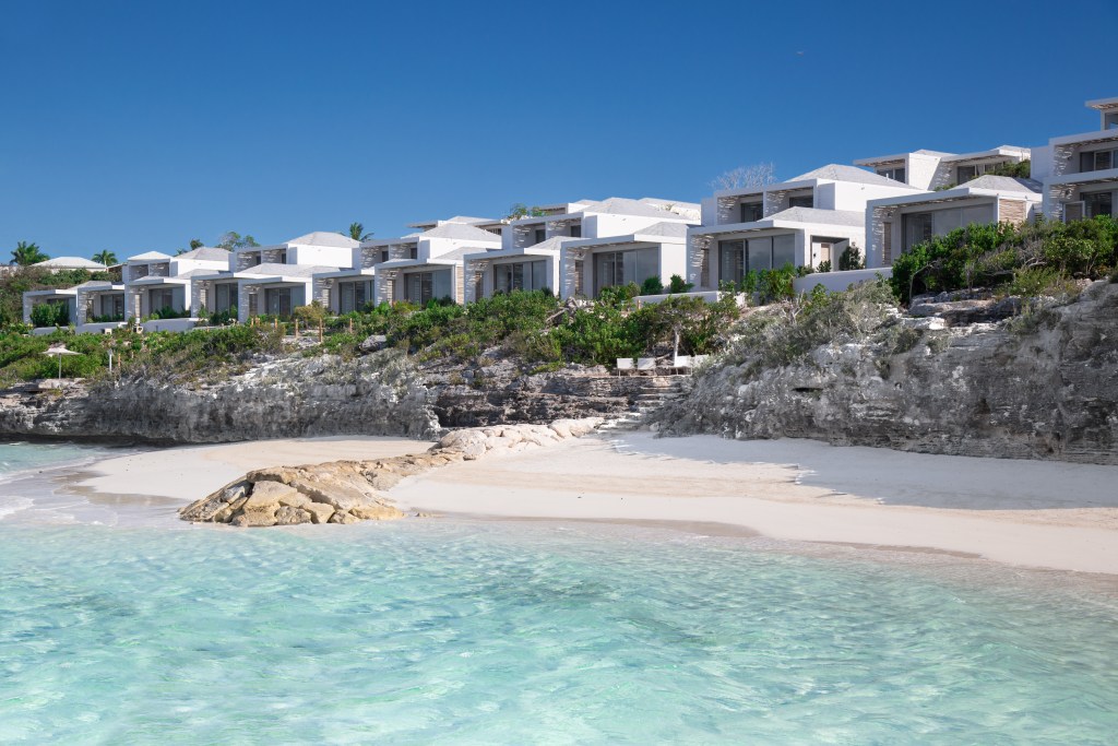 A small beach with crystal blue water and short cliffs running alongside white villas.