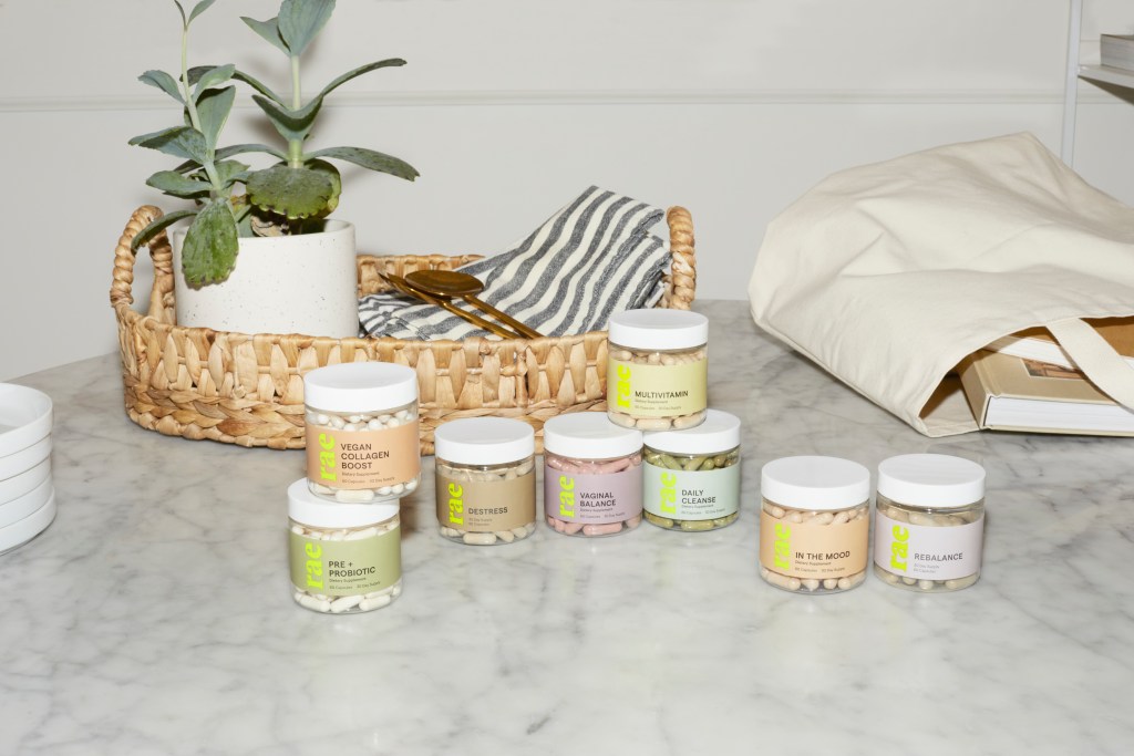 A white countertop with a shallow woven basket, a succulent plant, a tote bag of books and eight small jars of different Rae Wellness supplements.