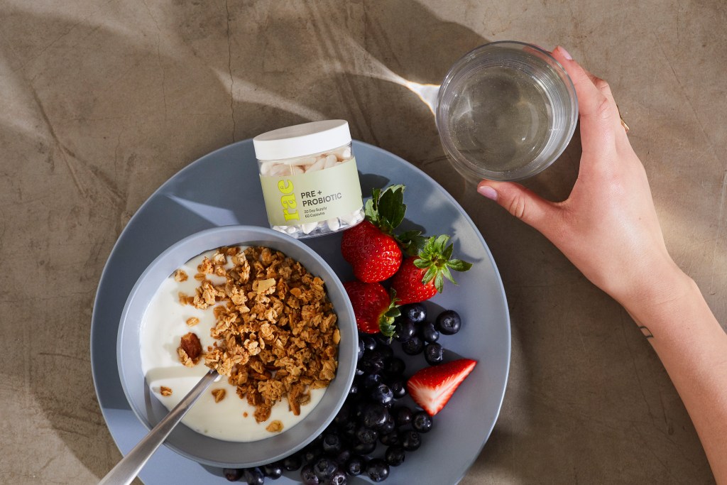 An overhead view of a woman's hand holding a glass of water next to a bowl of granola, berries and a jar of Rae Wellness supplements.