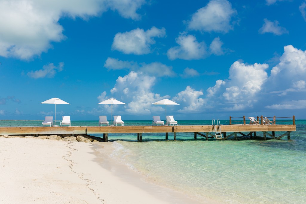 A long washed wood dock juts out from the sand into shallow crystal blue waters with a partly cloudy sky in the background.