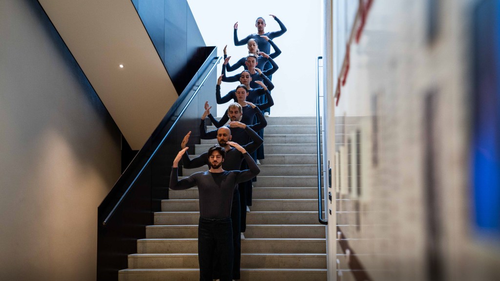A row of dancers wearing all black standing with arms raised in ascending order on a staircase.