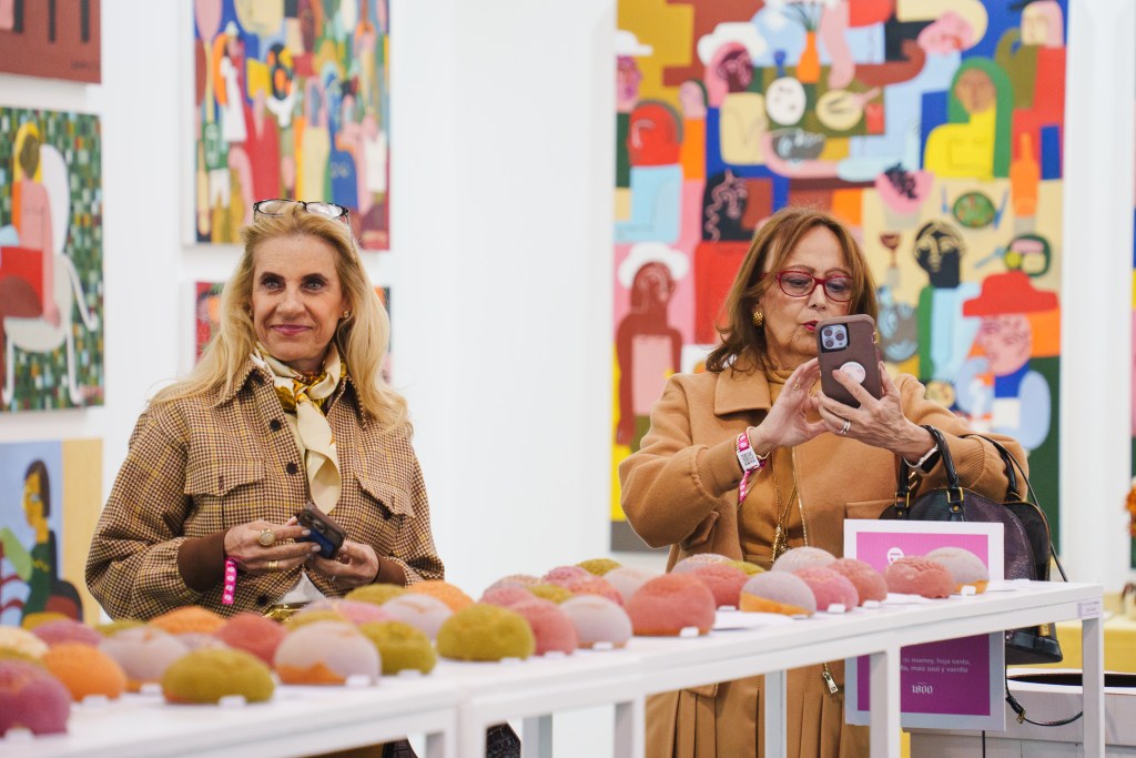 Two women looking at a table of coral, mustard and light lavendar sculptures at Zona Maco in Mexico City with large colorful paintings in the background.